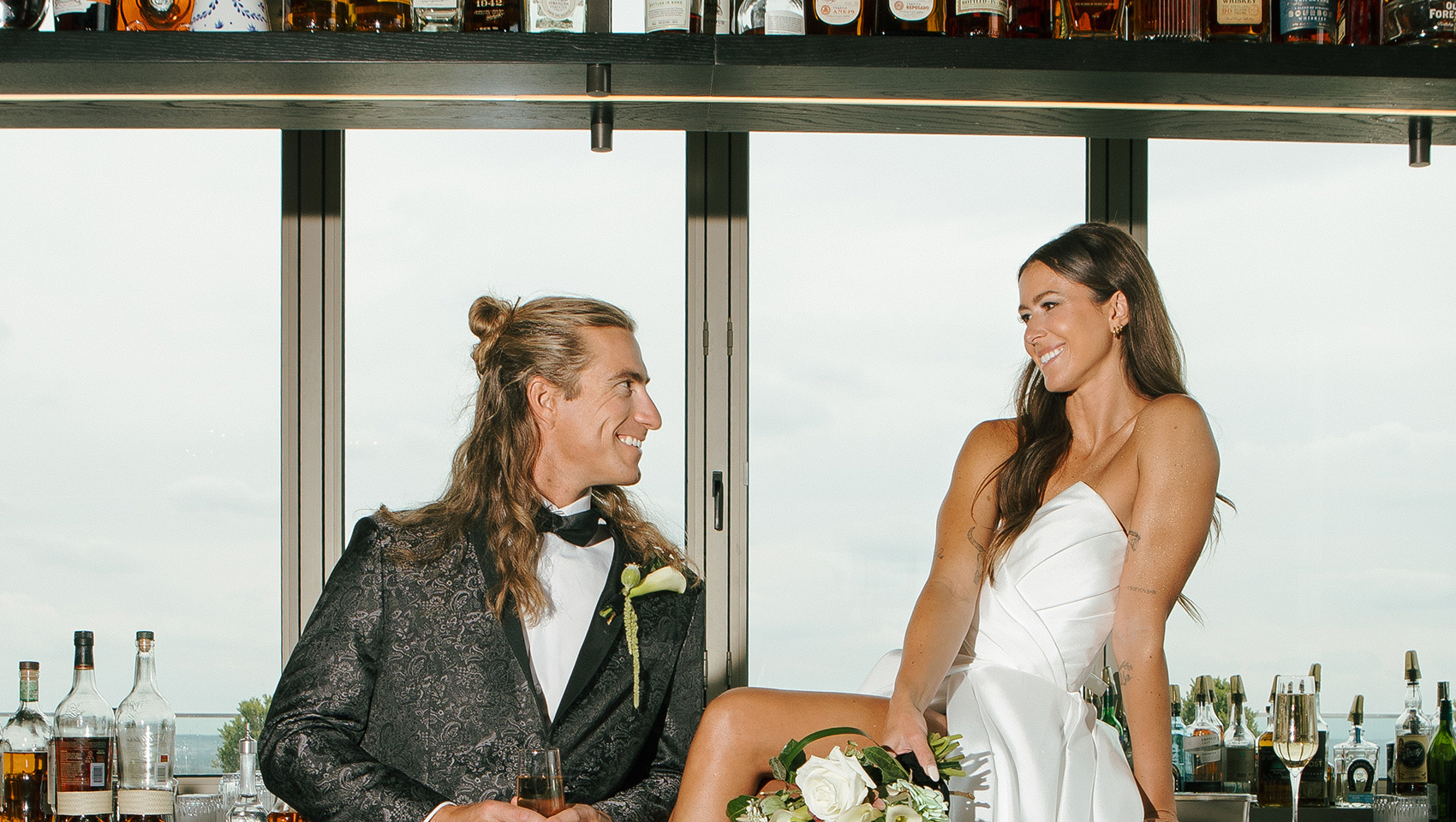 Bride and groom at halo bar. Bride sits on bar with bouquet and groom leans on bar holding a glass of champagne